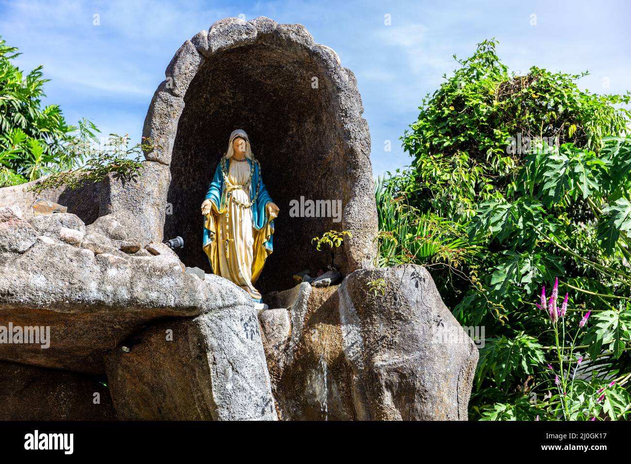Statua della Vergine Maria in una cappella rupestre fuori dalla chiesa cattolica di Sant’Anna a Baie Ste Anne, Praslin, Seychelles, con vegetazione tropicale Foto Stock
