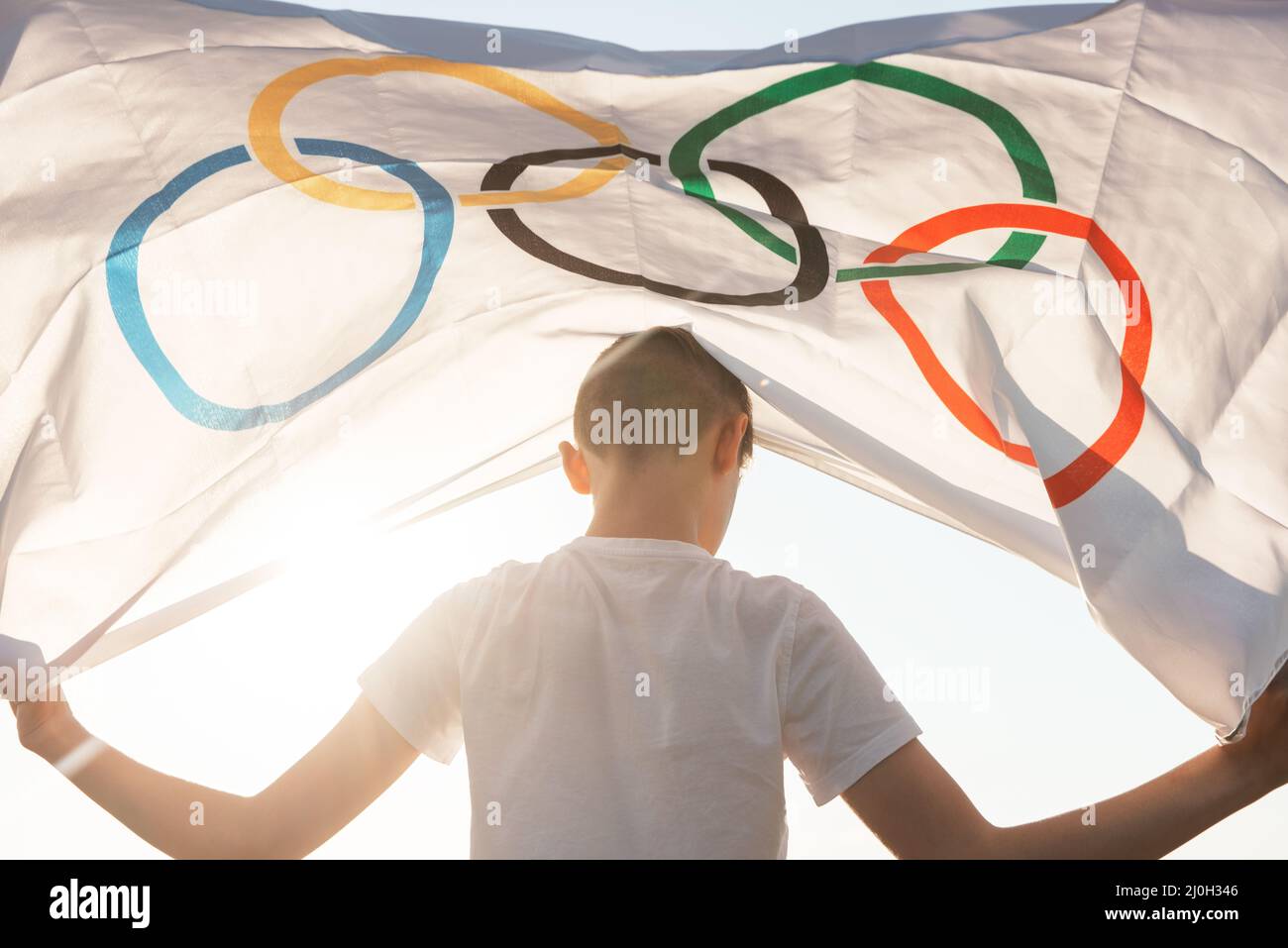 Il ragazzo biondo che sventola la bandiera dei Giochi Olimpici all'aperto sopra il cielo blu sulla riva del fiume Foto Stock