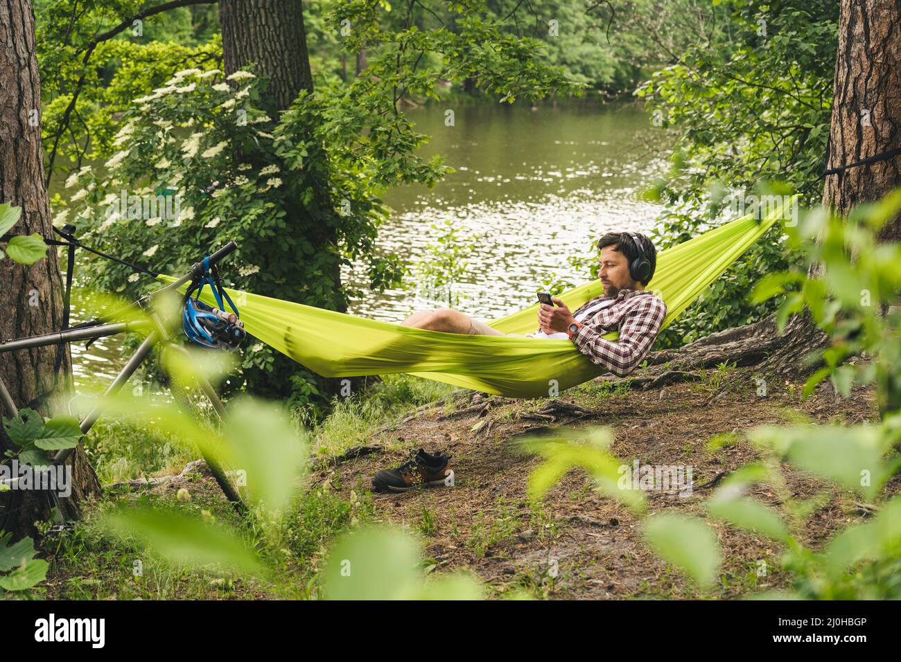 L'uomo viaggia in bicicletta, rilassandosi in amaca verde, navigando su Internet su smartphone, ascoltando musica sulle cuffie nella foresta vicina Foto Stock