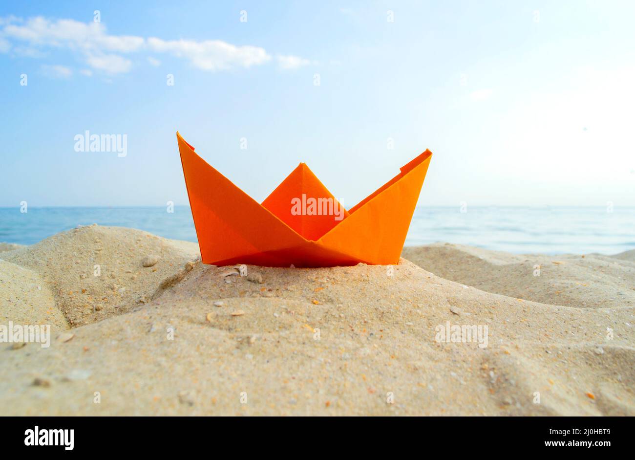 Barca di carta arancione brillante in sabbia gialla sullo sfondo delle onde del mare e del cielo blu Foto Stock