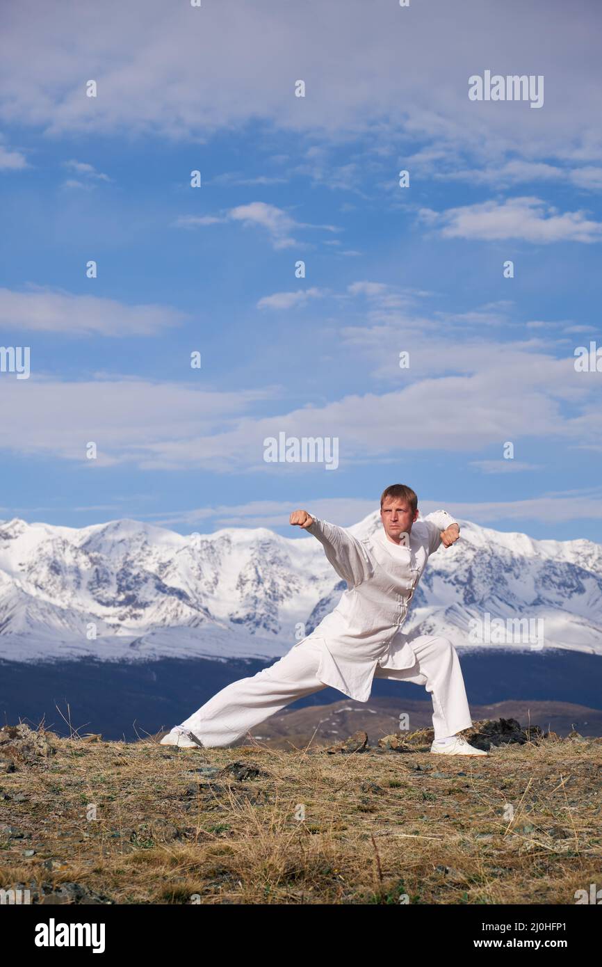 Wushu maestro in uno sport bianco uniforme di allenamento kungfu in natura sullo sfondo di montagne innevate. Foto Stock