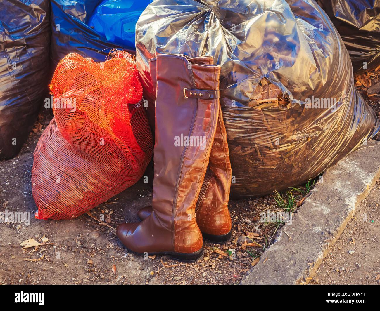 Stivali in pelle da donna in piedi vicino sacchi di rifiuti riempiti con fogliame asciutto autunno. Smaltimento di cose inutili. Raccolta rifiuti su strada Foto Stock