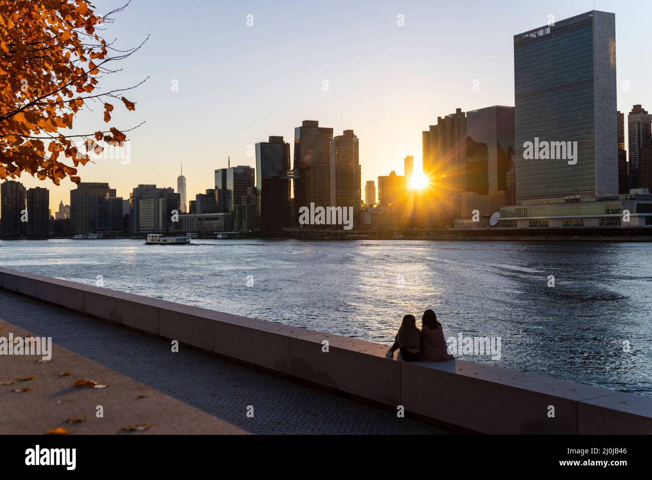 Il sole tramonta tra il grattacielo Midtown Manhattan oltre l'East River il 2021 novembre a New City. Due giovani donne siedono sul muro di Franklin D. Roo Foto Stock