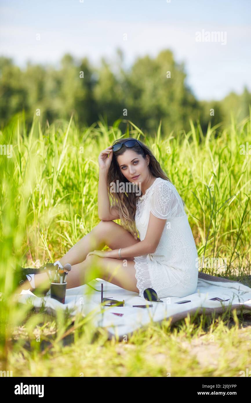 Giovane donna bionda in abito bianco e occhiali da sole è seduta su una coperta in erba alta. Foto Stock