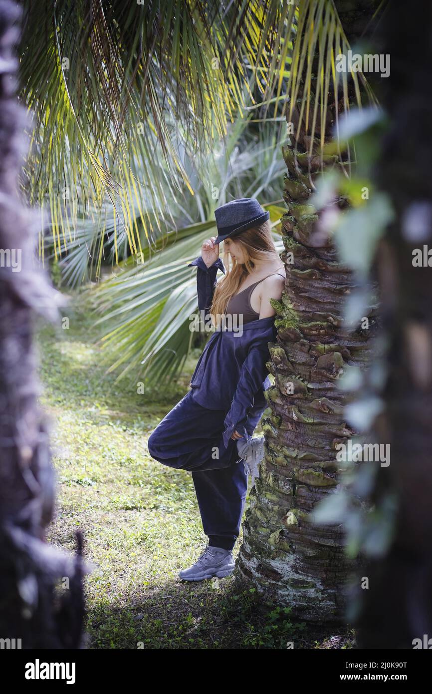 Una ragazza vestita casualmente sta tenendo la visiera di un cappello e appoggiandosi su una palma in un parco sud Foto Stock