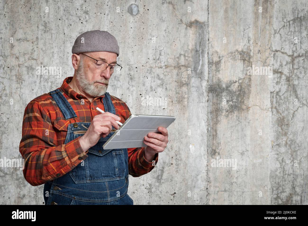 un uomo anziano sicuro che indossa occhiali, una camicia in flanella e tute sta prendendo appunti o schizzi su un tablet digitale Foto Stock