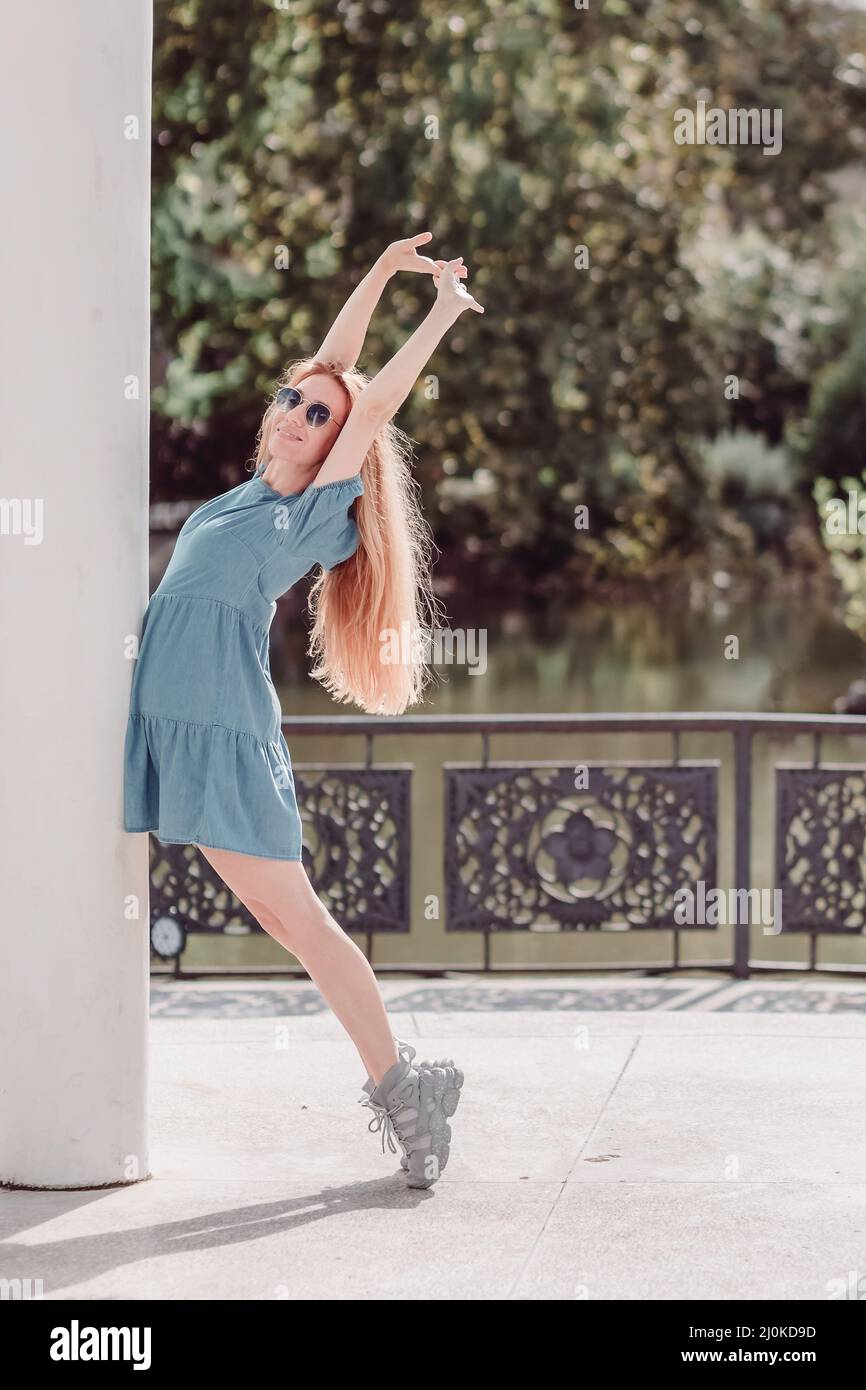 Una ragazza sorridente in un abito corto sta posando appoggiandosi su una colonna in piedi sulle sue dita dei piedi e sollevando le mani contro il backgrou Foto Stock