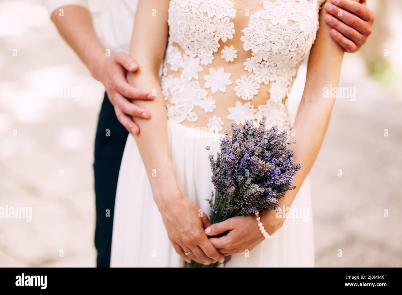 Lo sposo abbraccia da dietro in un vestito bianco di pizzo con un bouquet di lavanda nelle sue mani. Primo piano Foto Stock