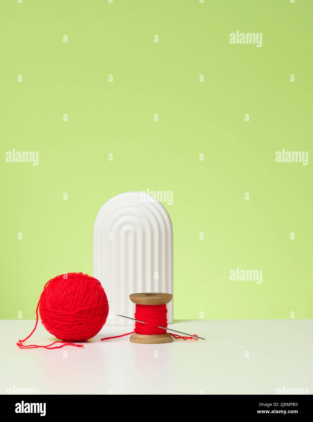 Palla rossa con filo di lana e grande ago su tavola bianca, sfondo verde Foto Stock