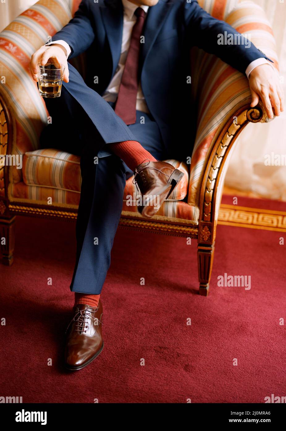 Le gambe di un uomo in pantaloni blu, calzini rossi e scarpe marroni seduti nella sedia che tiene il vetro in una mano Foto Stock