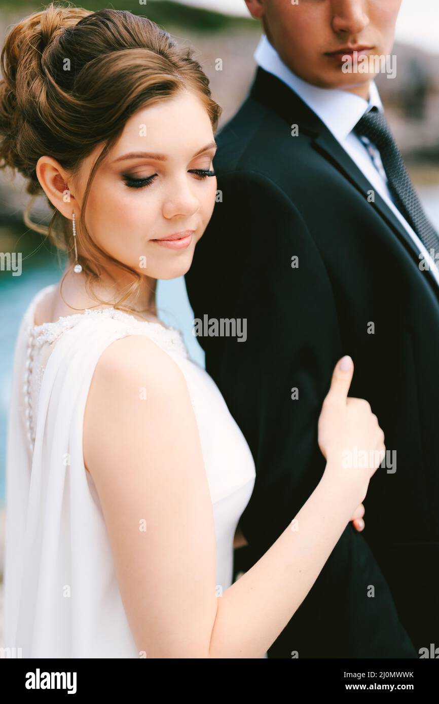 Sposa con acconciature alte in un abito bianco tiene il braccio dello sposo in una giacca nera e camicia bianca. Primo piano Foto Stock