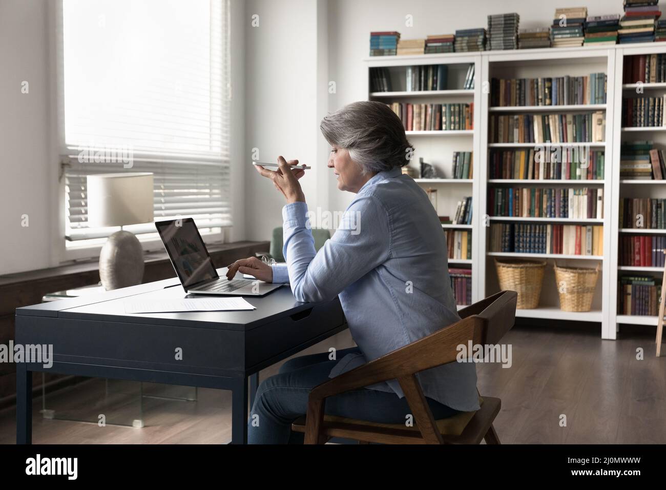 La donna d'affari anziana si siede alla scrivania utilizza lo smartphone parla con il vivavoce Foto Stock