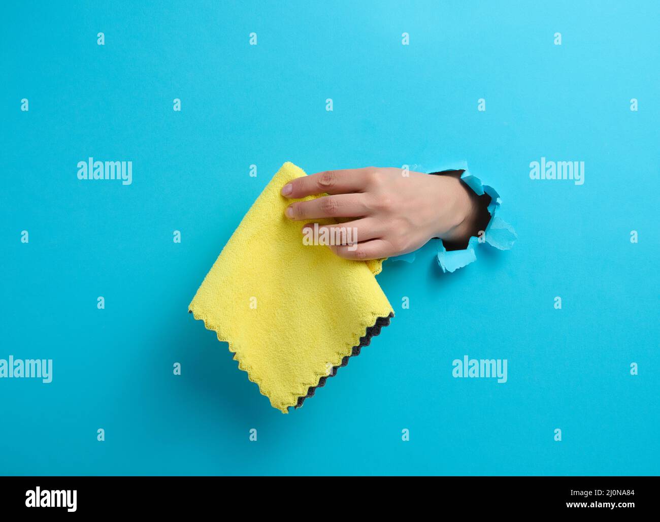 Mano femmina che sporge da un foro strappato in uno sfondo di carta blu e che tiene uno straccio asciutto Foto Stock