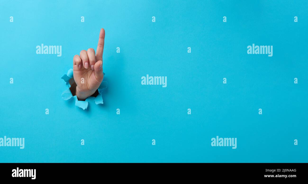 Mano femmina che sporge da un foro strappato su uno sfondo di carta blu, gesto di attenzione. Luogo per un'iscrizione, banner Foto Stock