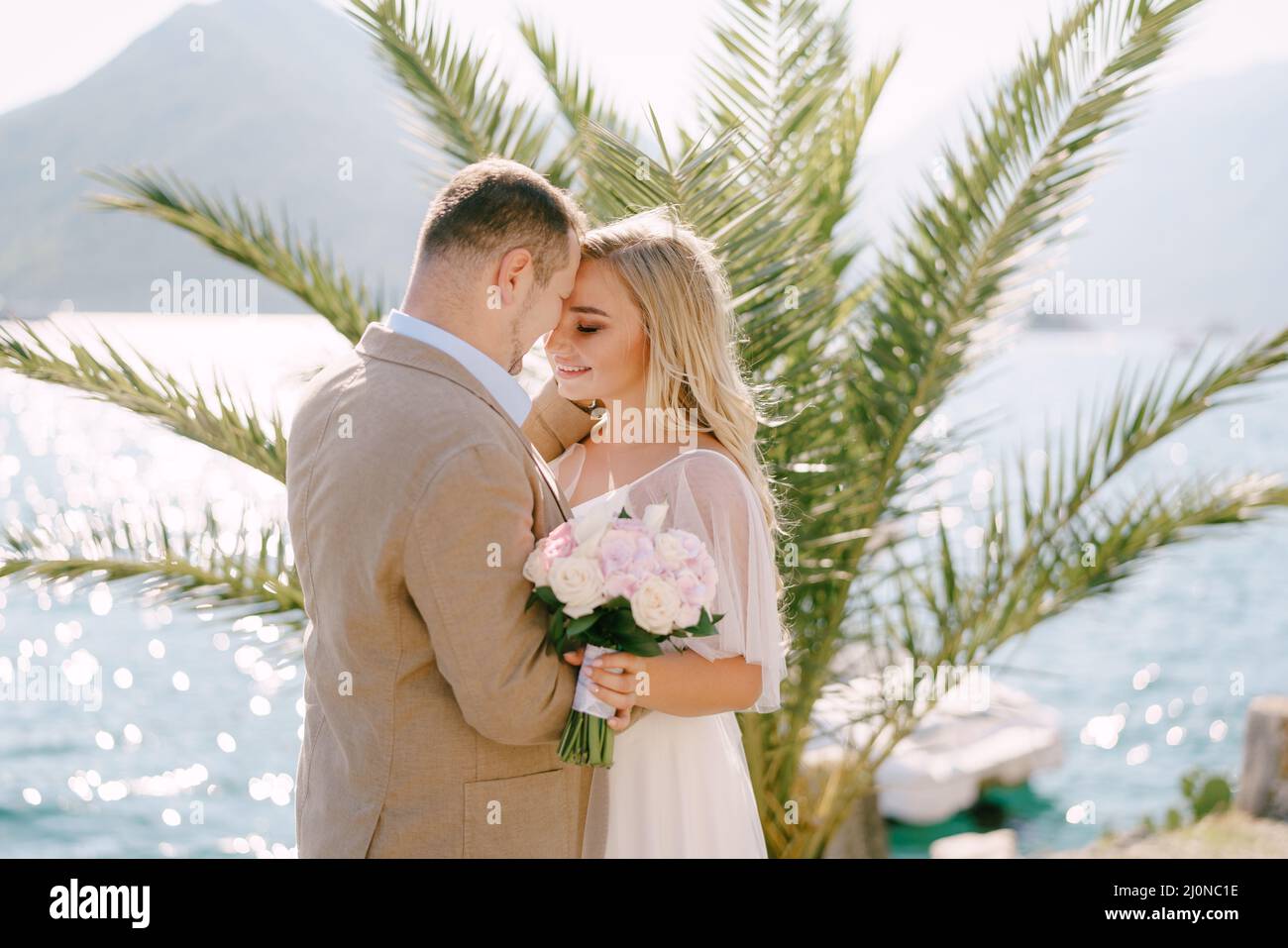 Lo sposo e la sposa sorridente con un bouquet stand sul molo sullo sfondo di una palma e teneramente abbraccio Foto Stock