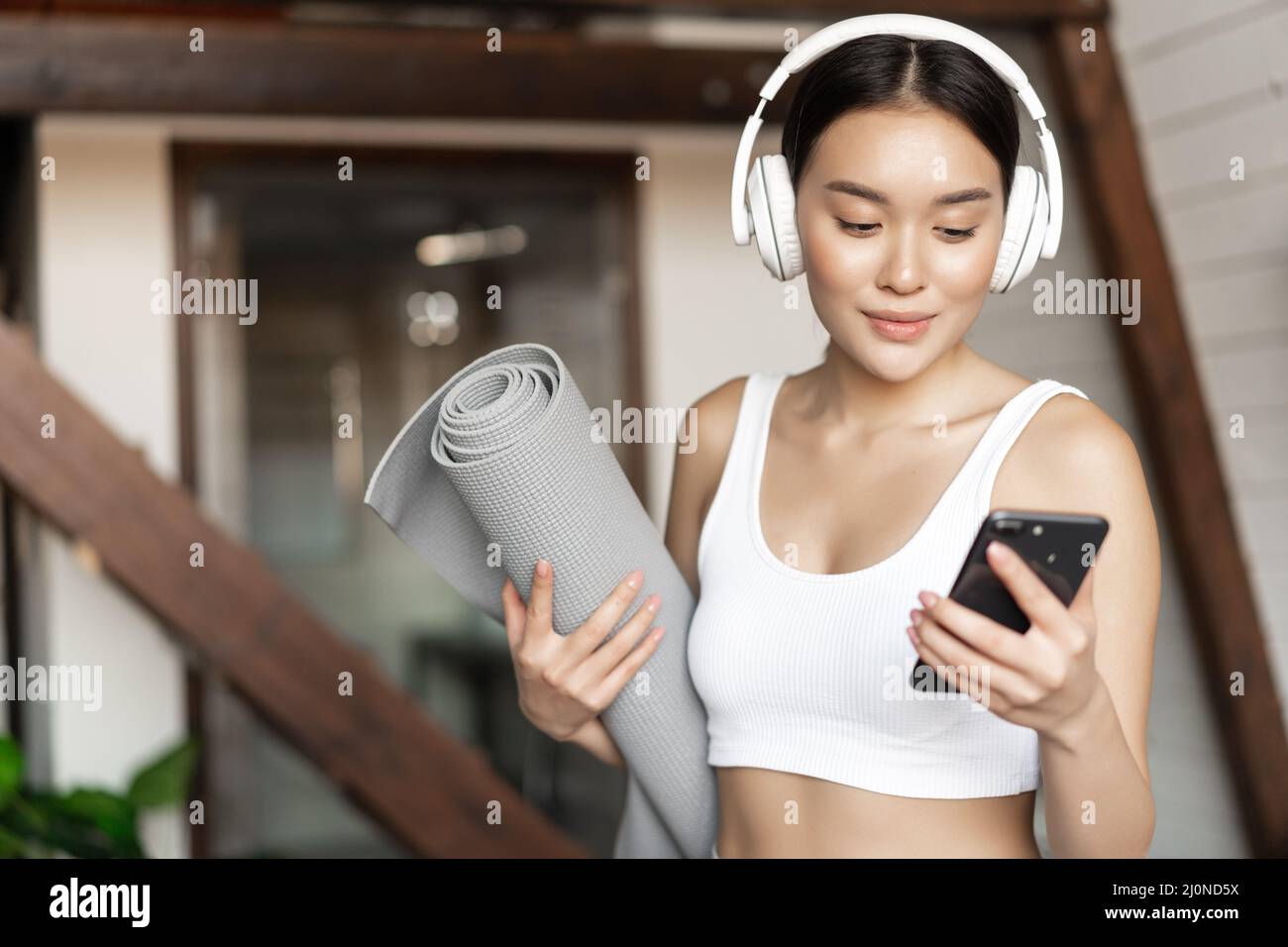 Giovane donna asiatica in piedi a casa in cuffie, ascoltare musica e andare in allenamento sul tappetino, praticare yoga o meditare in l Foto Stock