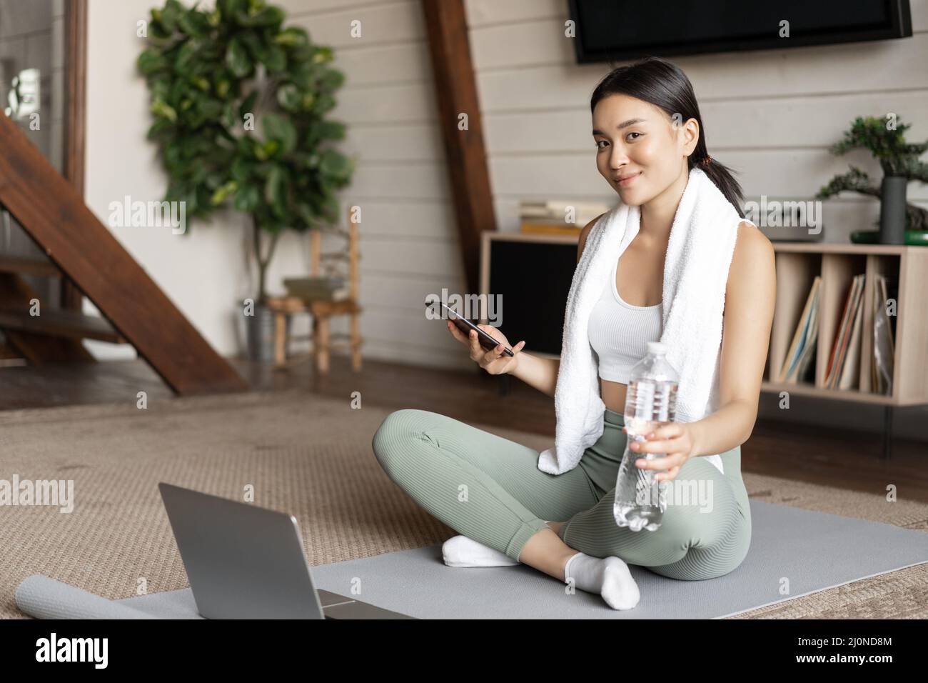 Ragazza asiatica in abbigliamento fitness seduta a casa sul tappetino, riposo dopo l'allenamento, controllo dell'applicazione sportiva del telefono cellulare Foto Stock