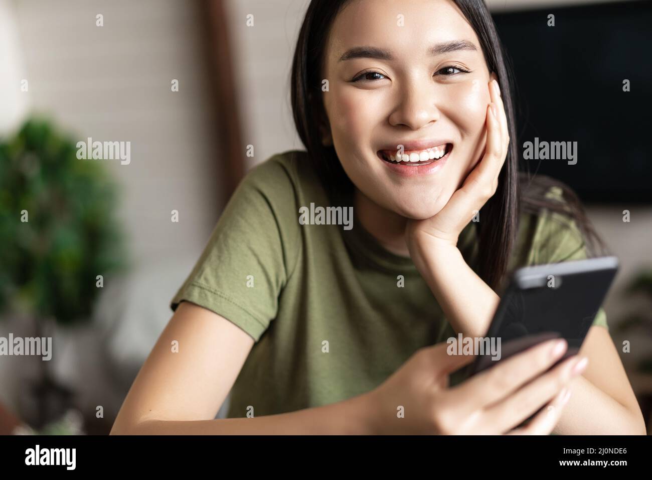 Sorridendo donna asiatica usando il telefono cellulare da casa, facendo ordine, consegnare smth a casa sua Foto Stock