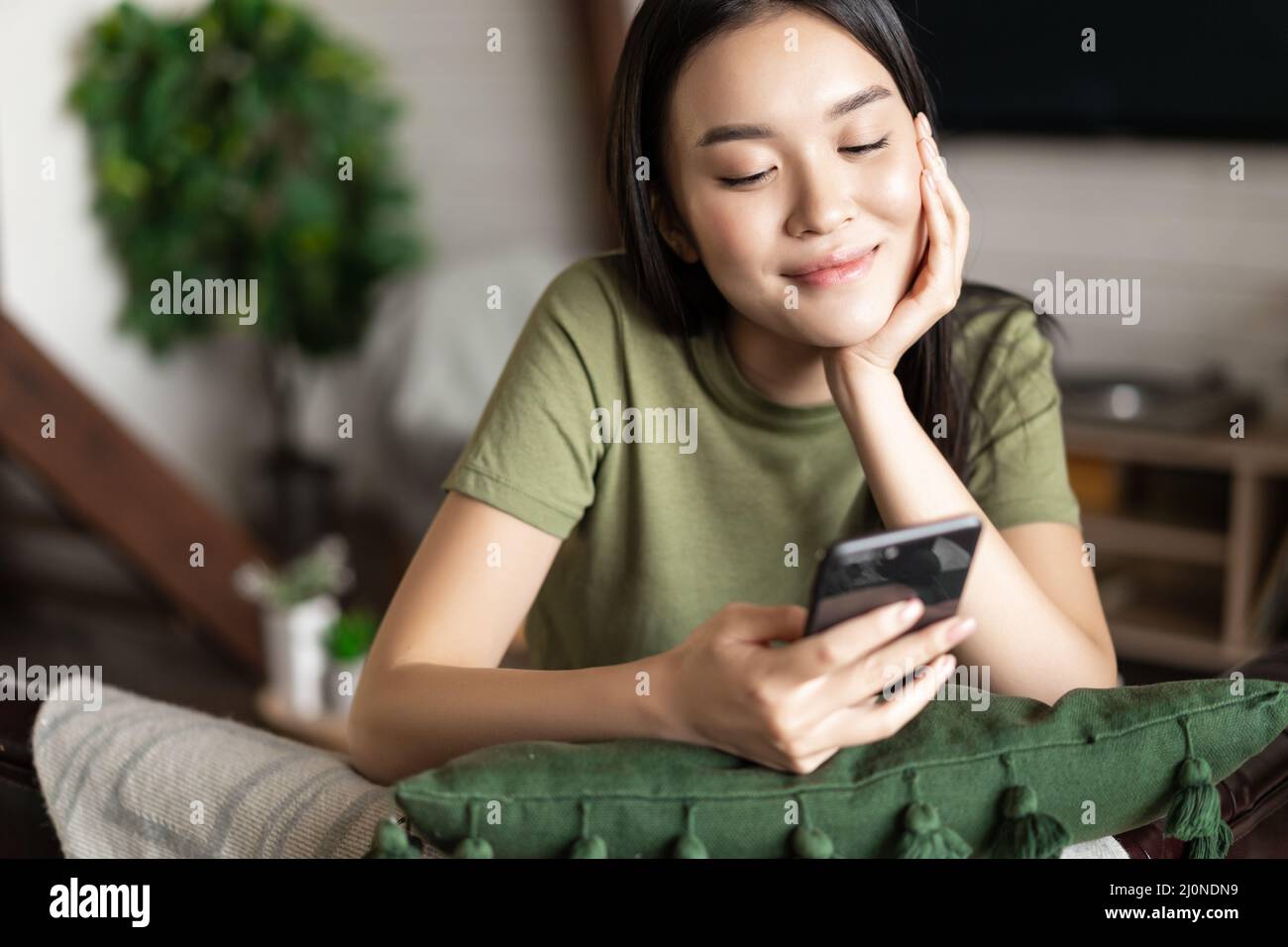 Ragazza coreana sorridente che guarda il telefono cellulare, seduto a casa in una t-shirt casual Foto Stock