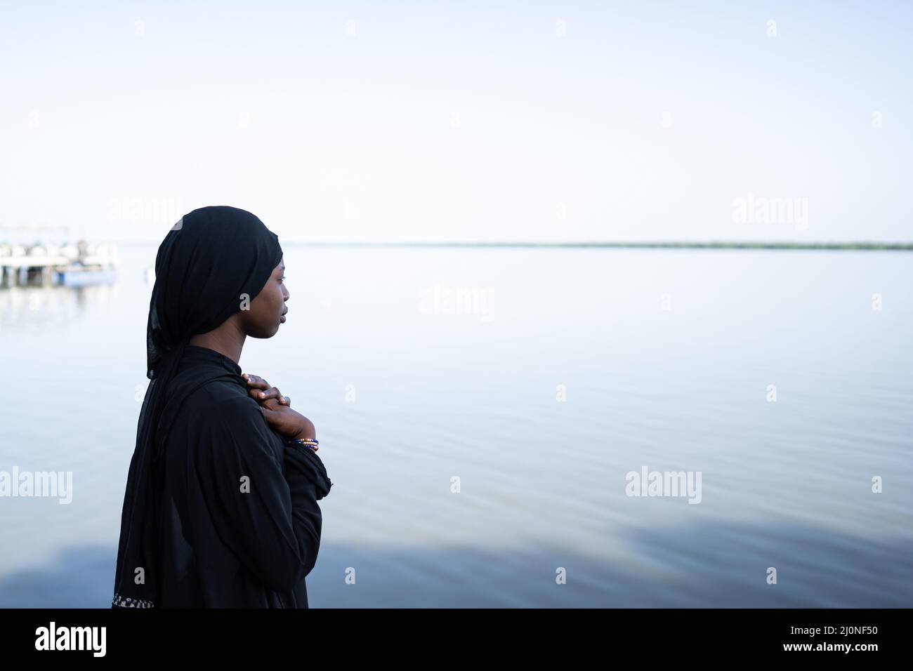 Ragazza africana solitaria e travagliata, vestita tutta con una scialle nera e una lunga veste, di fronte al mare, mani incrociate sul petto in un gesto ansioso; Lon Foto Stock