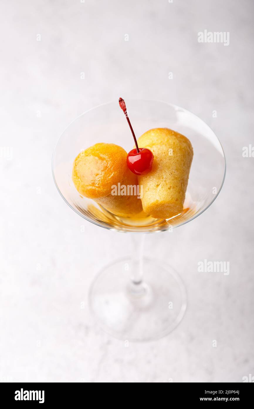 Rum baba napoletano (o baba au rum) in un bicchiere di martini con una ciliegia da cocktail su sfondo grigio. Piccole torte di lievito imbevute di sciroppo di rum. Tradizione Foto Stock