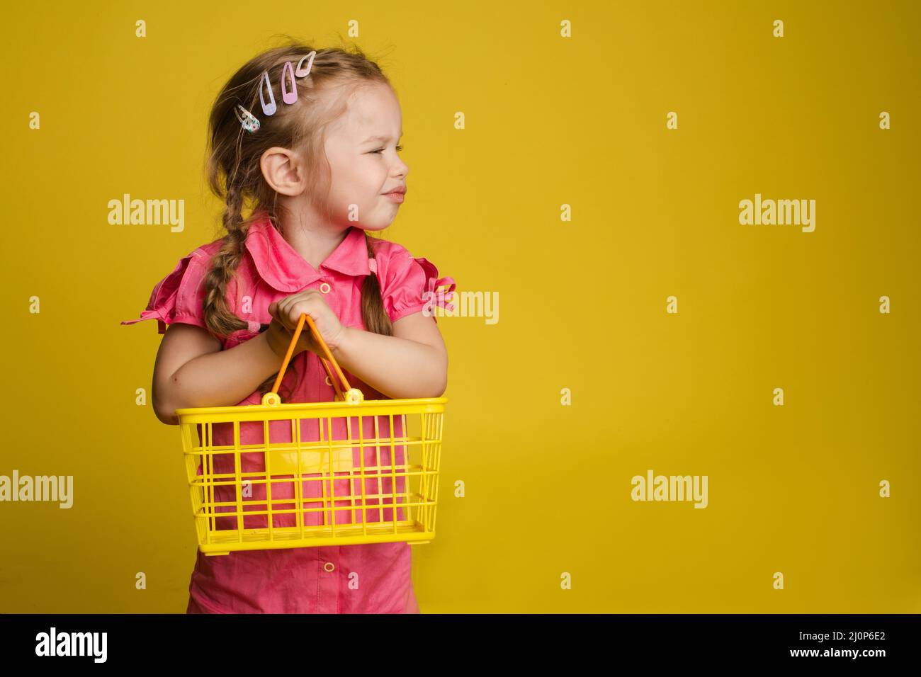 Cunting piccola ragazza carina che tiene piccolo cestino che posa isolato a sfondo giallo studio Foto Stock