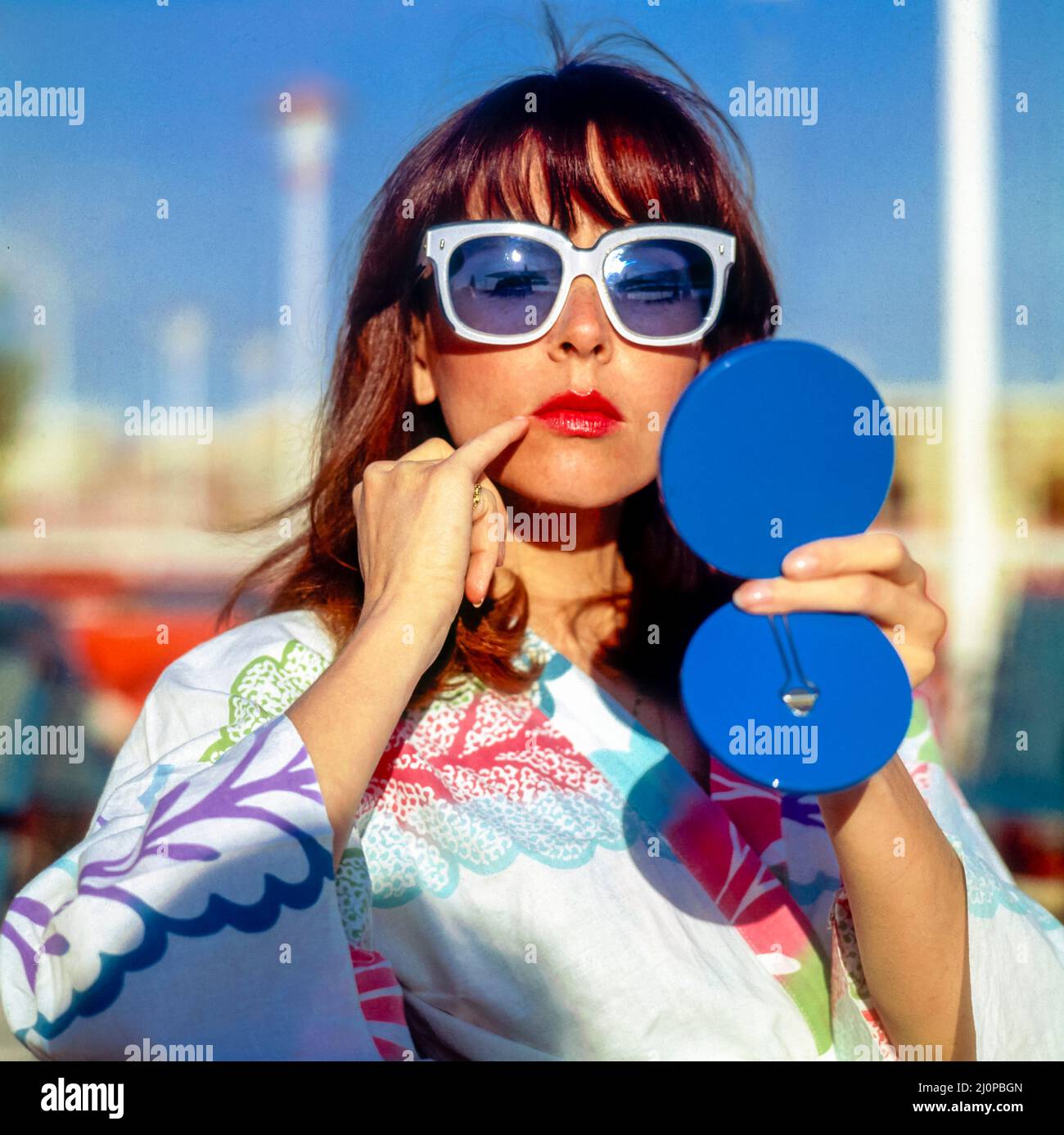 Vintage Italy 1970s, graziosa donna con occhiali da sole bianchi che guarda  in uno specchio blu compatto che controlla il suo trucco, spiaggia di  Kursaal, Lido di Ostia, Europa Foto stock -