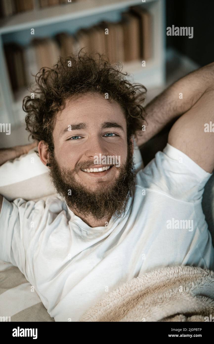 Un bel ragazzo con occhi blu, capelli ricci, una barba e un ampio sorriso si trova sotto la copertura del suo letto. Vista dall'alto. Primo piano verticale. Foto Stock