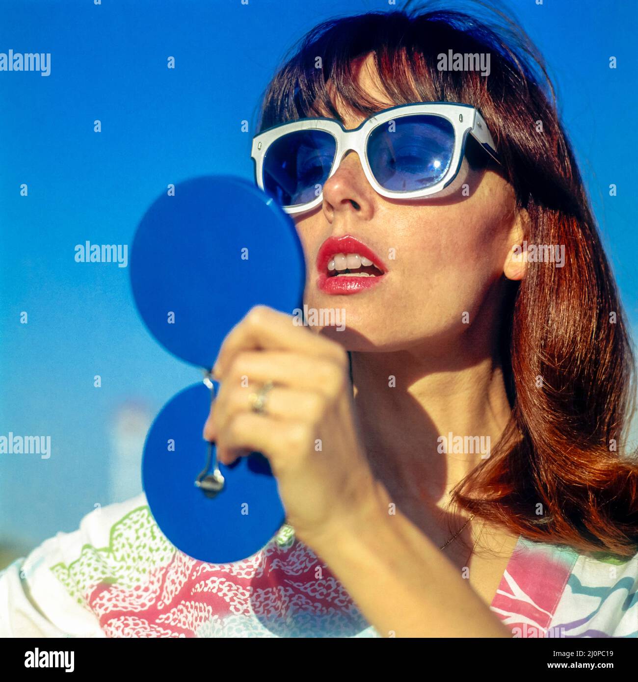 Vintage Italy 1970s, graziosa donna con occhiali da sole bianchi che guarda  in uno specchio blu compatto che controlla il suo trucco, spiaggia di  Kursaal, Lido di Ostia, Europa Foto stock -