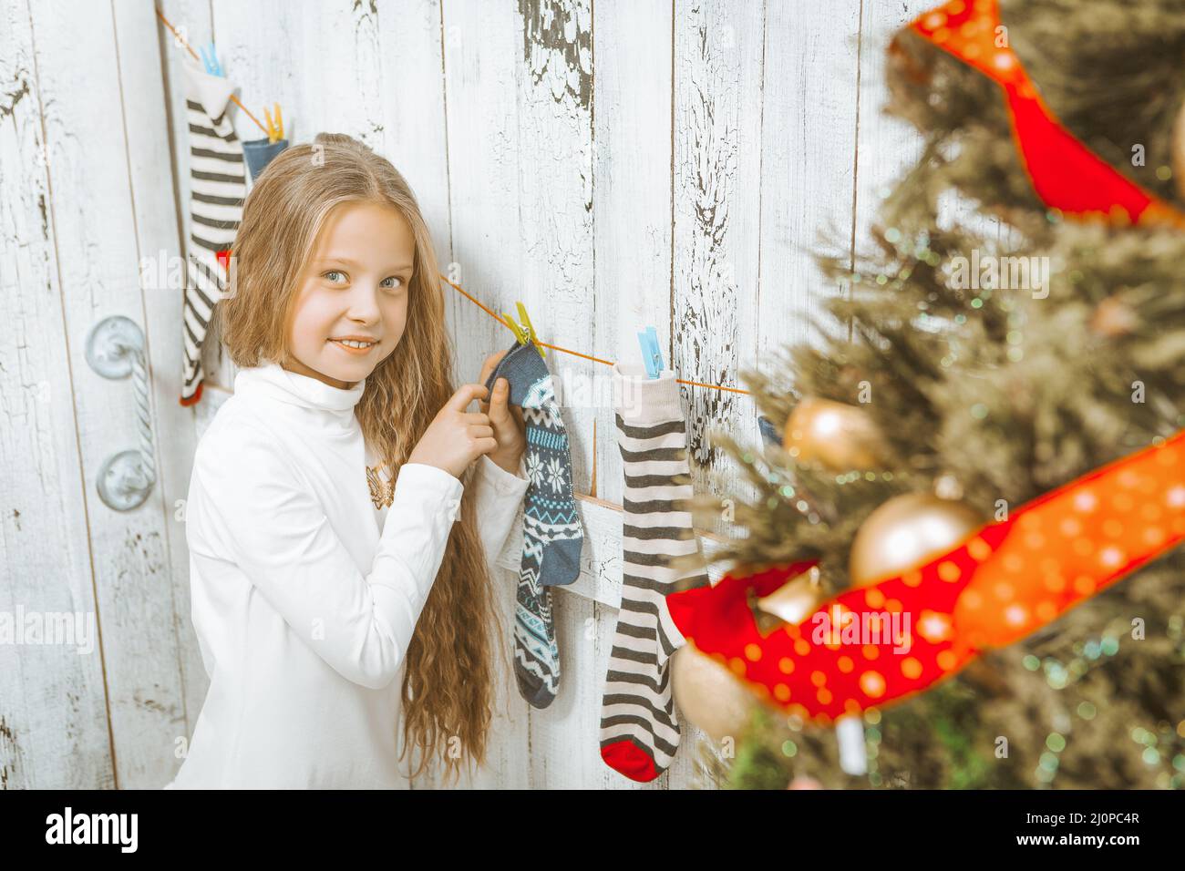 Ragazza caucasica carina in un ponticello bianco con capelli biondi lunghi appende calze di Natale. Calze di Natale. Sfondo di legno bianco. Cl Foto Stock