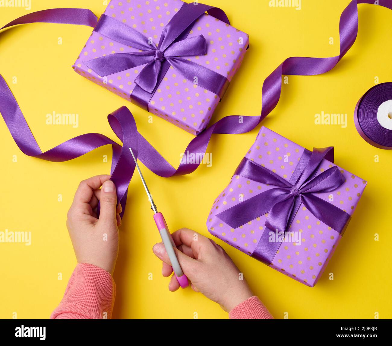 Le mani femminili stanno tagliando un nastro viola di seta con le forbici, vicino alle scatole del regalo. Sfondo giallo, vista dall'alto Foto Stock