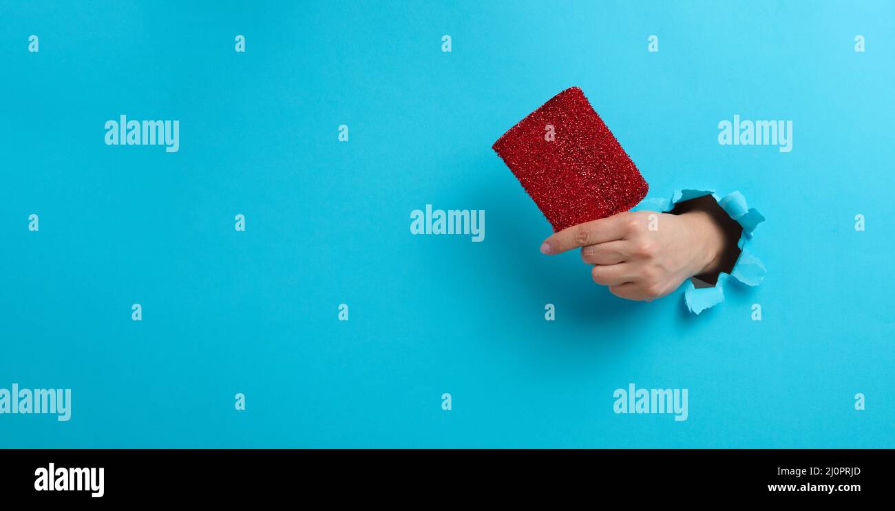 La mano femmina tiene una spugna rossa da cucina. Parte del corpo che sporge da un foro strappato su uno sfondo di carta blu Foto Stock