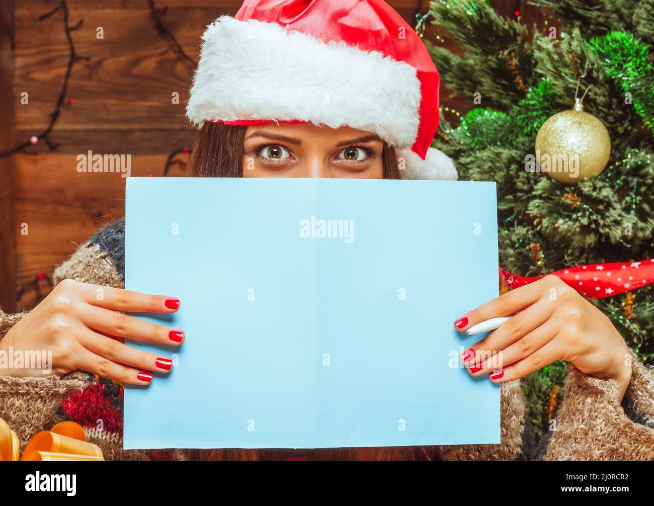 Modello caucasico femmina in un cappello Santa coperto la parte inferiore del suo viso con un foglio di carta bluastro, ha penna in mano. Let Foto Stock