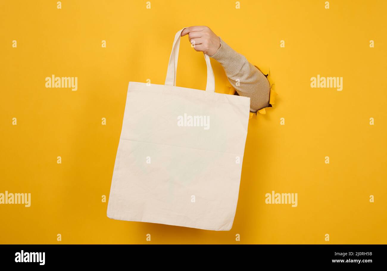 La mano femmina tiene un sacchetto bianco vuoto di tessuto su uno sfondo giallo, una parte del corpo sporge da un foro strappato in un bac di carta Foto Stock