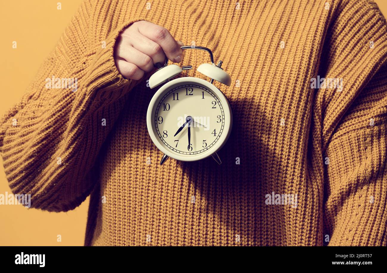 Sveglia rotonda in mano di una donna, il tempo è la metà delle sette del mattino. Donna in maglione arancione Foto Stock