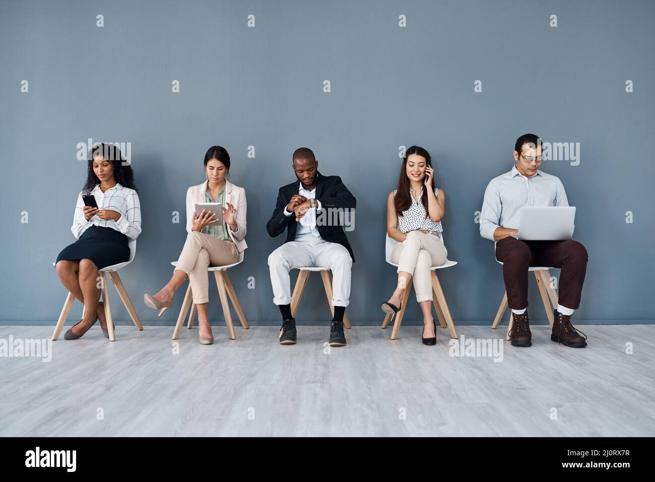 In attesa del loro turno per un colpo all'avanzamento di carriera. Scatto di un gruppo di uomini d'affari che utilizzano dispositivi digitali mentre si siedono in linea contro un grigio Foto Stock