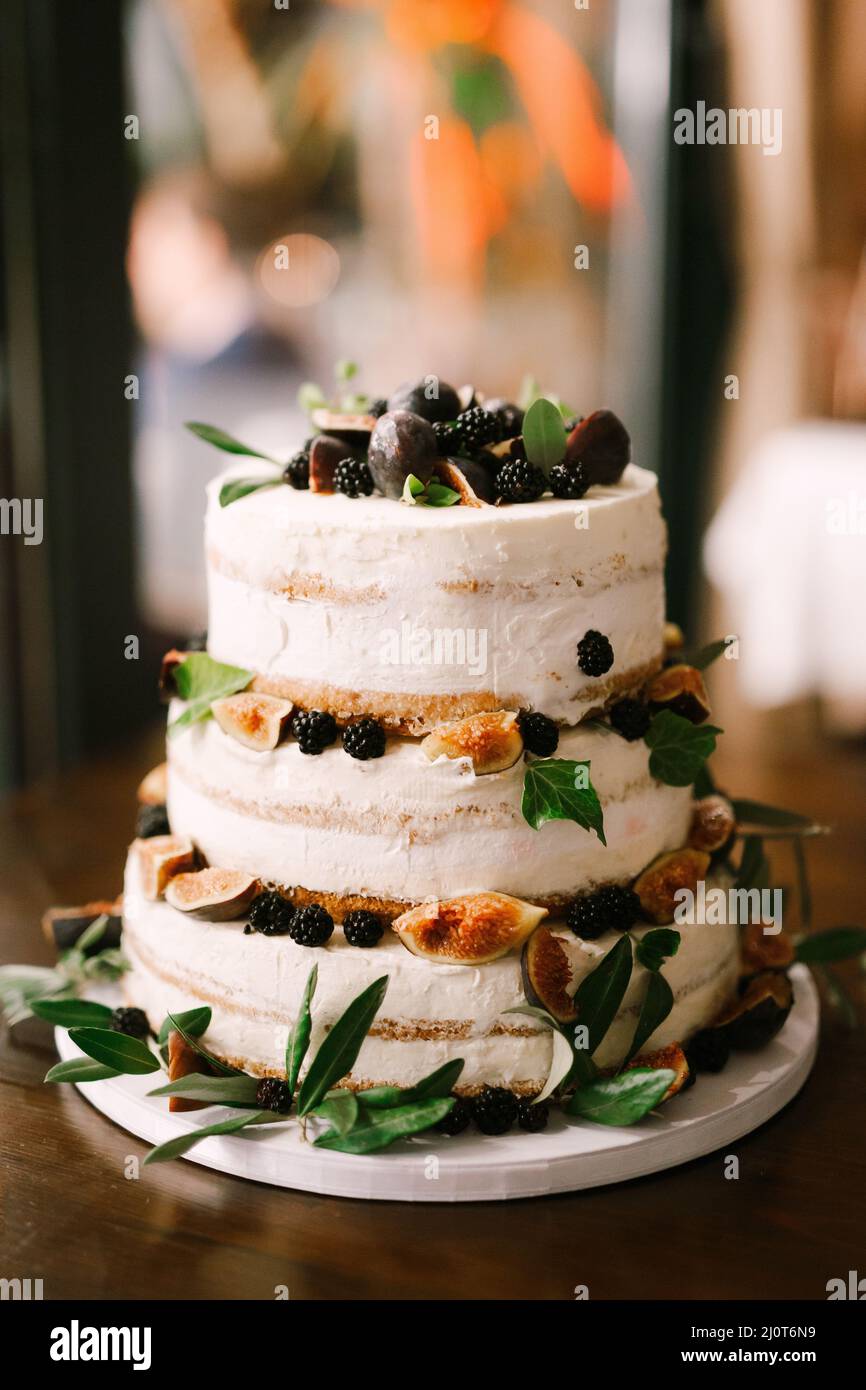 Torta nuziale a tre piani decorata con more, fichi, prugne e foglie verdi si erge sul tavolo Foto Stock