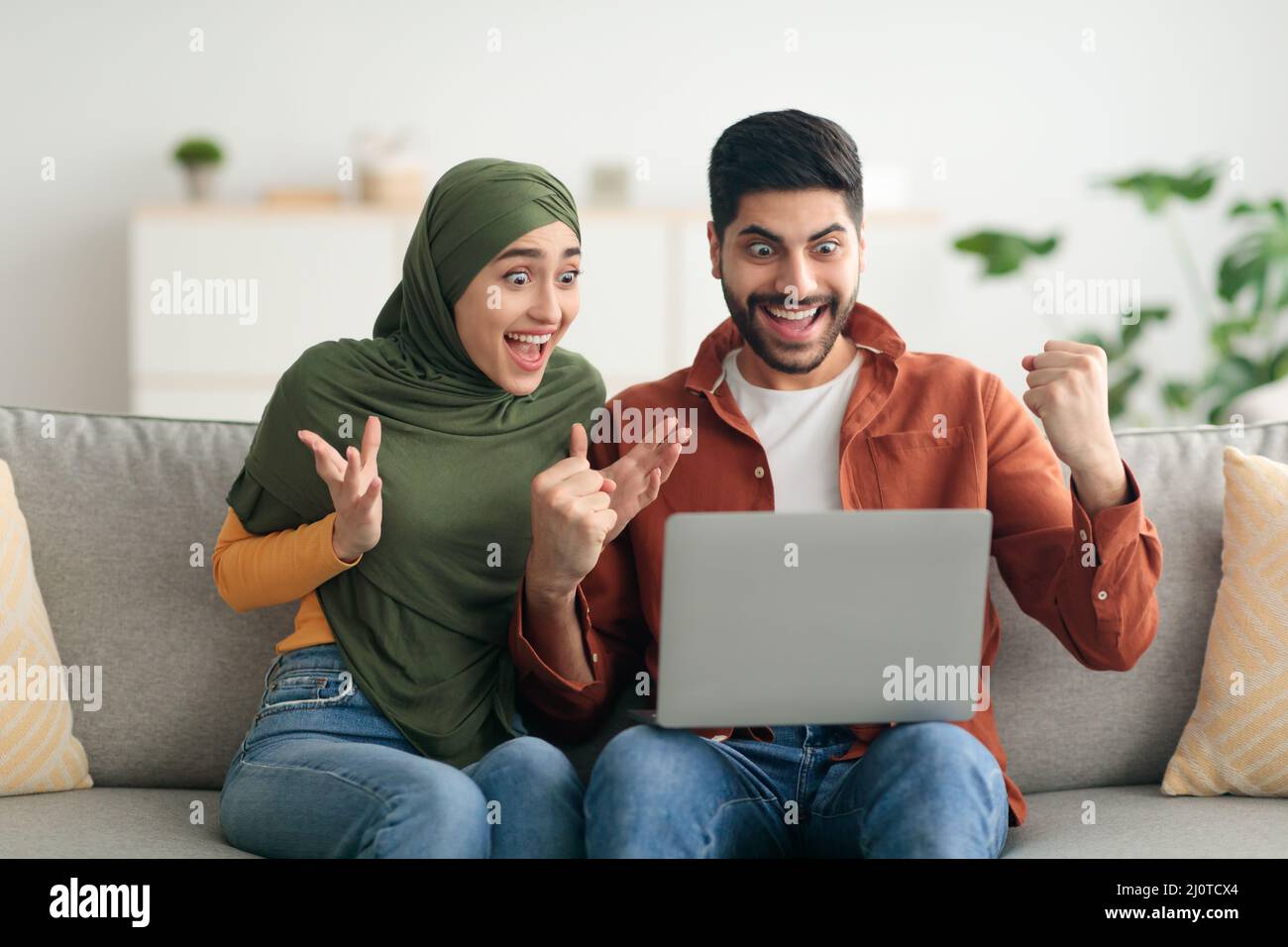 Joyful sposi musulmani usando il laptop che celebra la vittoria scuotendo i fists all'interno Foto Stock