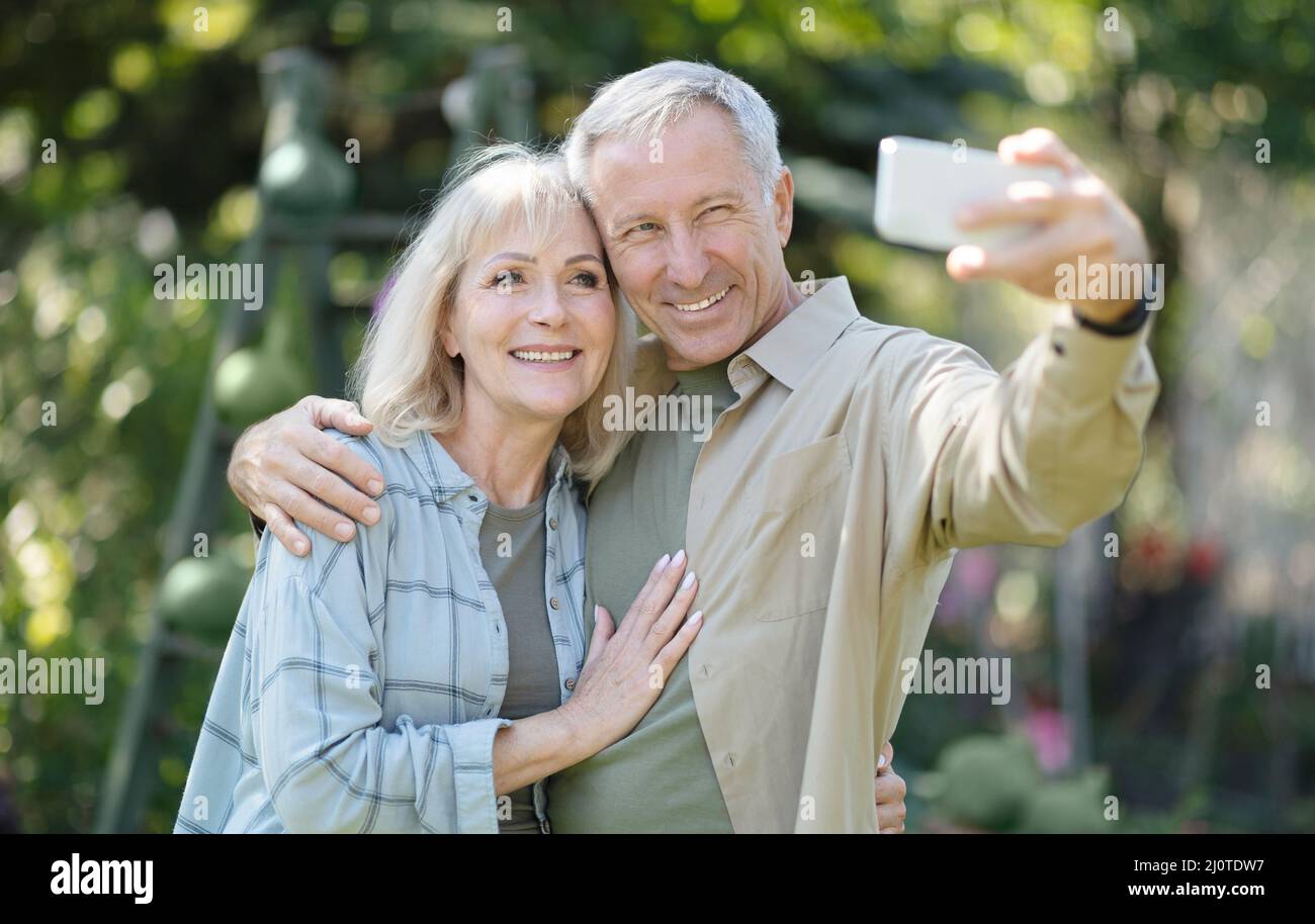 Coppia di anziani felici che trascorrono il tempo nel loro giardino insieme, prendendo selfie e sorridendo alla macchina fotografica del cellulare Foto Stock