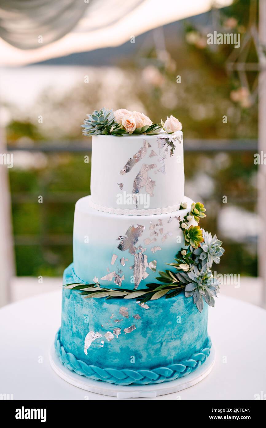 Torta nuziale bianca e blu con fiori e succulenti si erge su un tavolo bianco Foto Stock