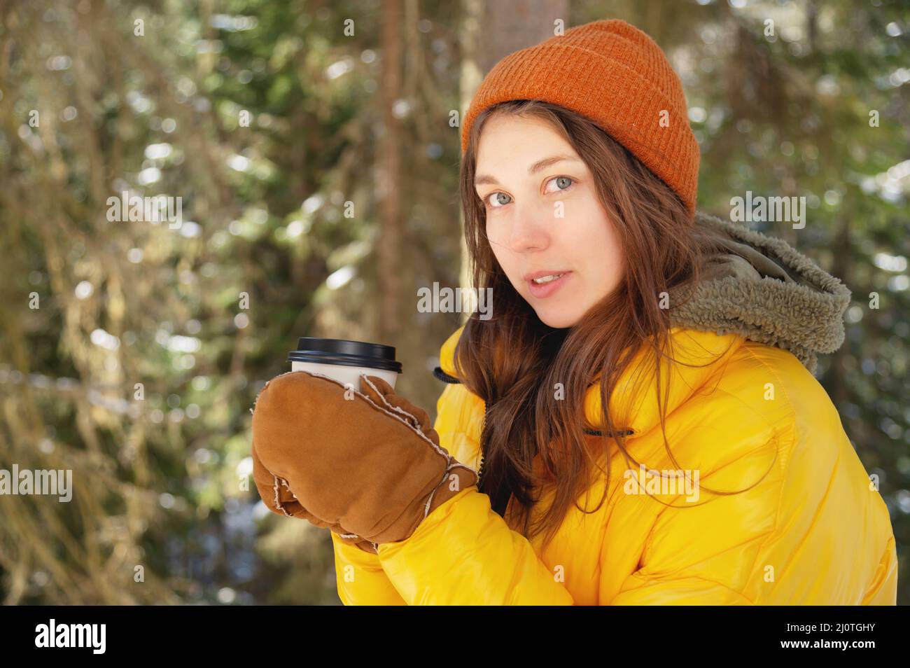 Bella caucasica positiva giovane donna in abiti invernali in un cappello con guanti e una giacca in giù si erge in un vino conifere Foto Stock