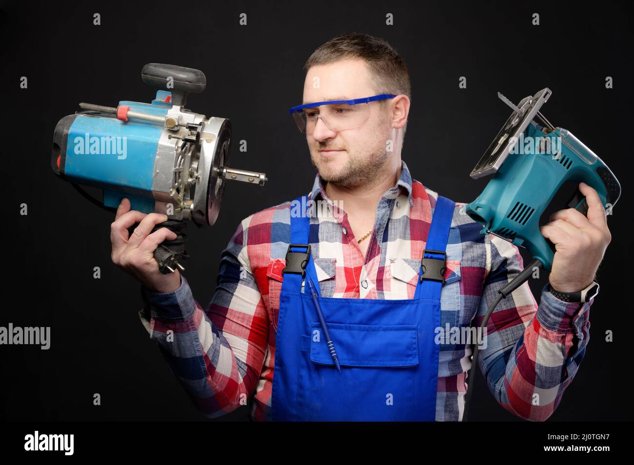 Un falegname professionale in uniforme e occhiali di protezione contiene un utensile elettrico, un seghetto alternativo e una fresa. Ritratto studio su nero Foto Stock
