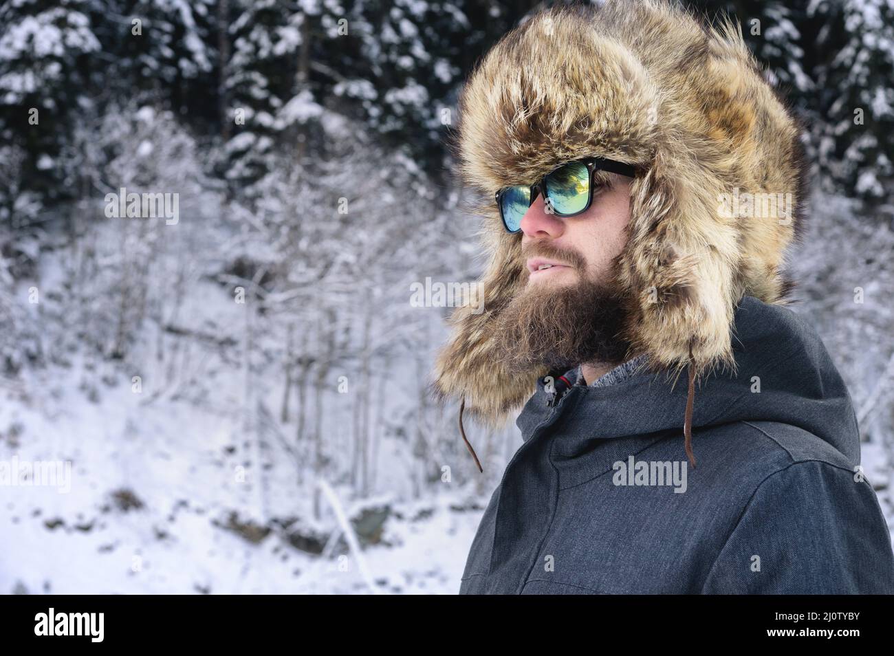 Ritratto di un giovane attraente uomo portico in un grande cappello di pelliccia e occhiali da sole contro la foresta innevata in montagna Foto Stock