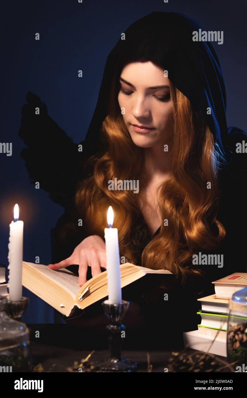 Medio primo piano attraente ragazza caucasica con capelli lunghi dorati in un cappuccio nero siede ad un tavolo e foglie attraverso un libro. Tuta w Foto Stock