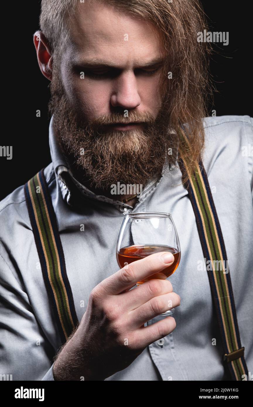Un uomo attraente con una barba lunga e un baffo seduto contro il muro sniffa il profumo di una bevanda alcolica in un Foto Stock