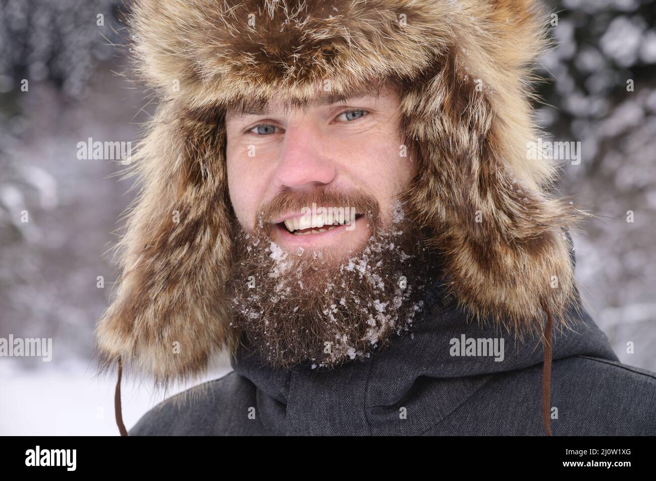 Ritratto di un uomo bearded in un cappello grande di pelliccia sullo sfondo di una foresta innevata. Uomo di manly sorridente nei boschi Foto Stock