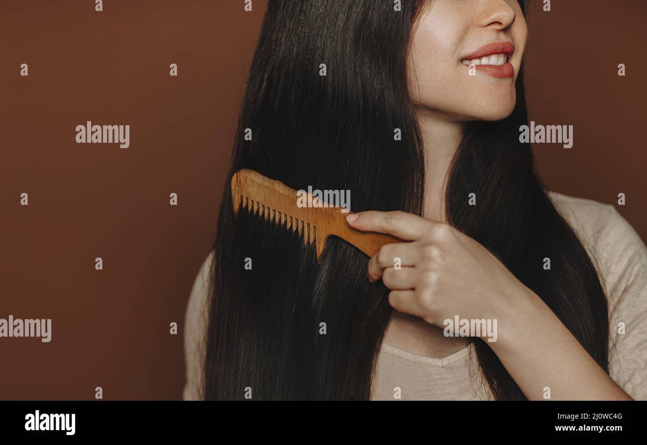 Giovane donna bruna felice pettinando i suoi capelli lunghi sani e sani e sorridendo alla macchina fotografica mentre si posa isolato su sfondo scuro Foto Stock