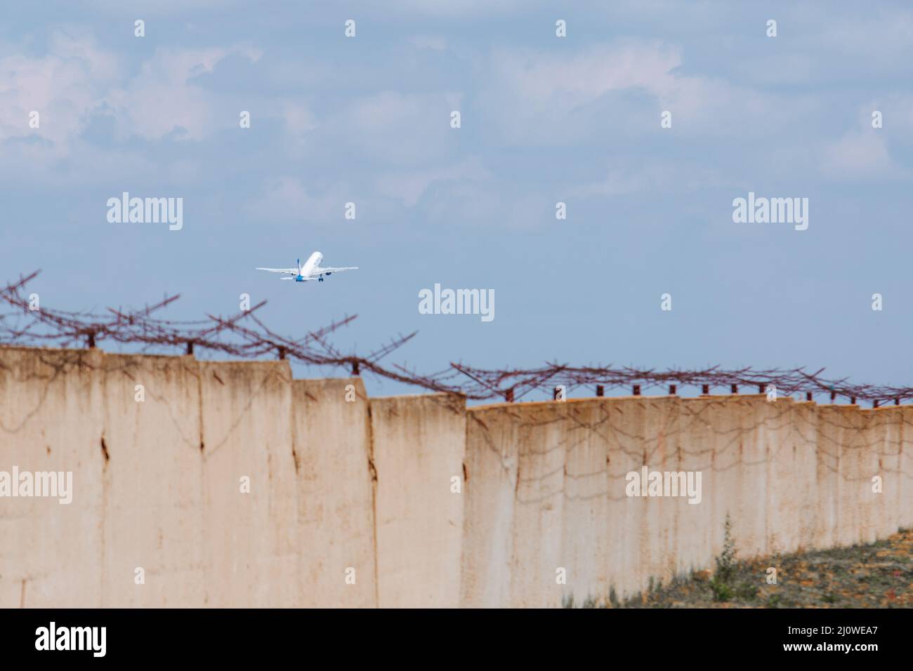 L'aereo nel cielo sullo sfondo di un muro di cemento con filo spinato. Evacuazione. Crimine, fuga. Foto Stock