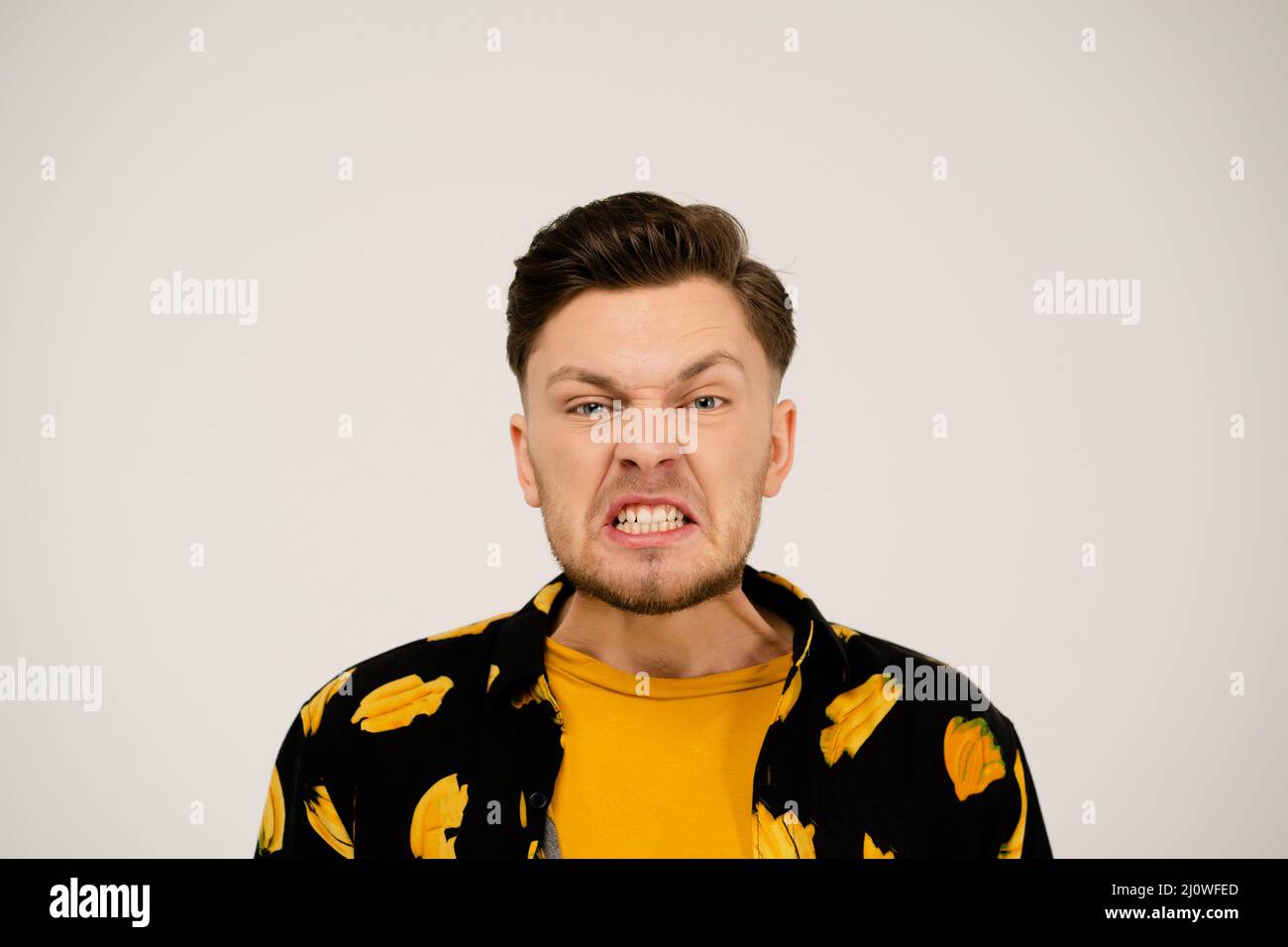 L'uomo in una T-shirt gialla e una maglietta divertente mostra un'emozione negativa per la fotocamera su uno sfondo bianco nello Studio. Ragazzo arrabbiato. Foto Stock