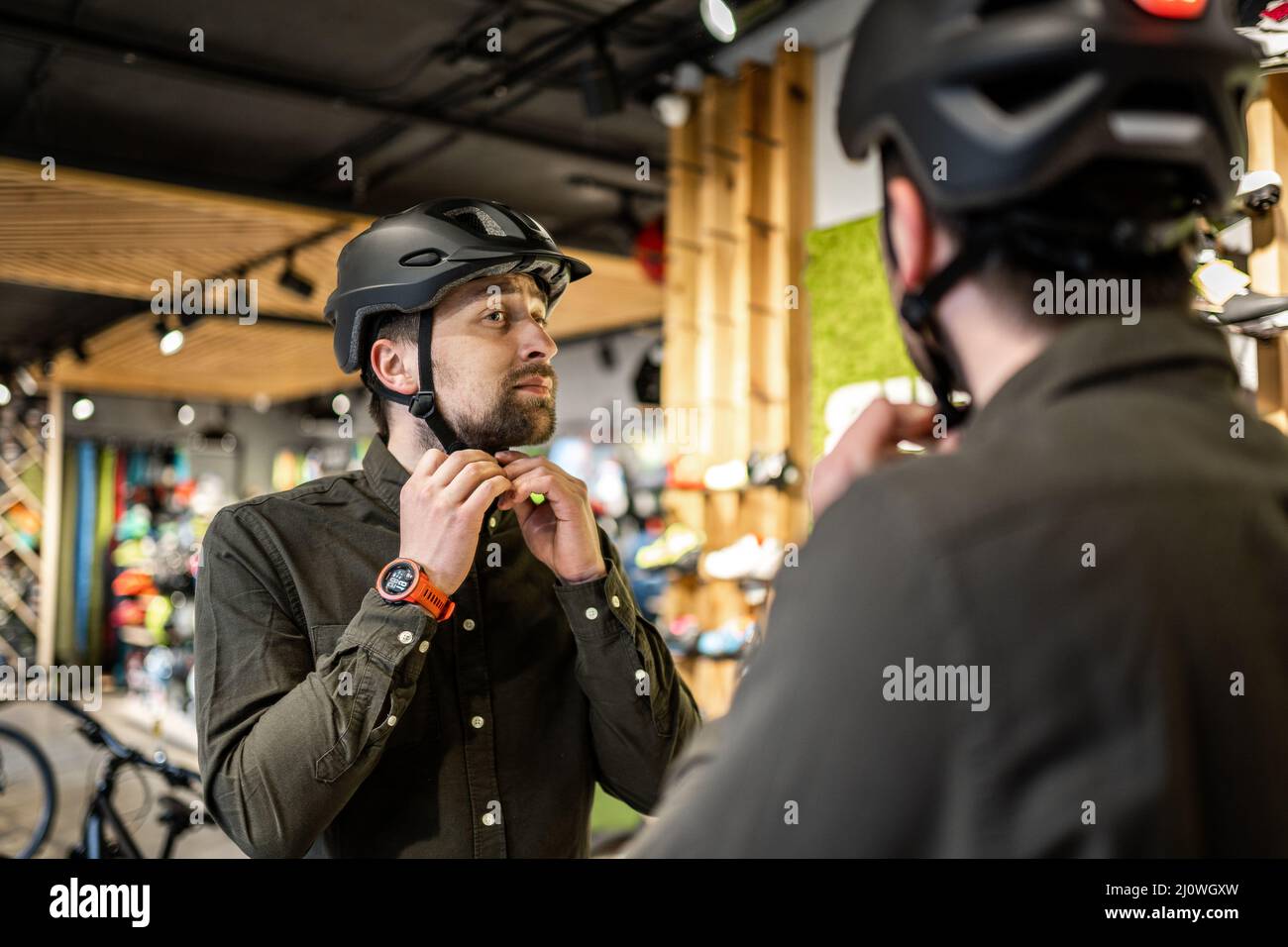 Il giovane uomo è venuto al negozio di biciclette. Sta misurando il casco. Il maschio sceglie il casco nel negozio di attrezzature sportive. Acquisto di nuovo Foto Stock